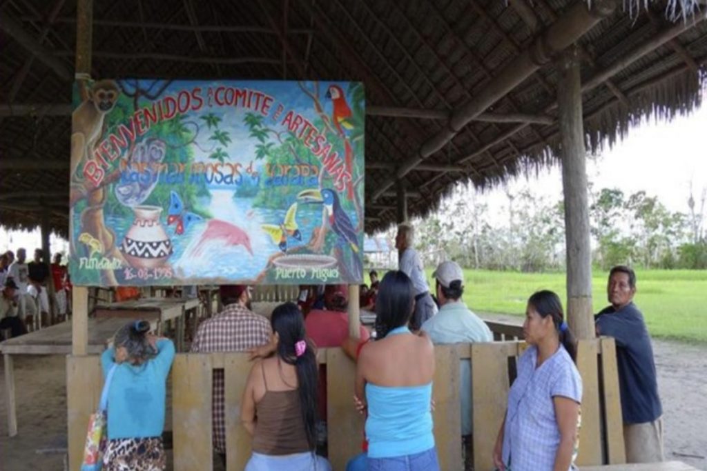 A colorful mural welcomes visitors to a handicraft committee, depicting jungle animals and flora. People are seated under a thatched roof structure, facing the mural.